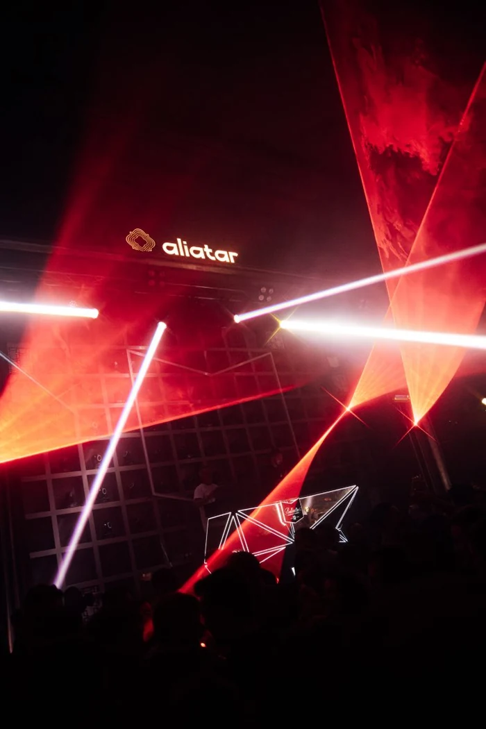 Láseres rojos cruzando la sala sobre la cabina, con el rótulo de Aliatar iluminado al fondo